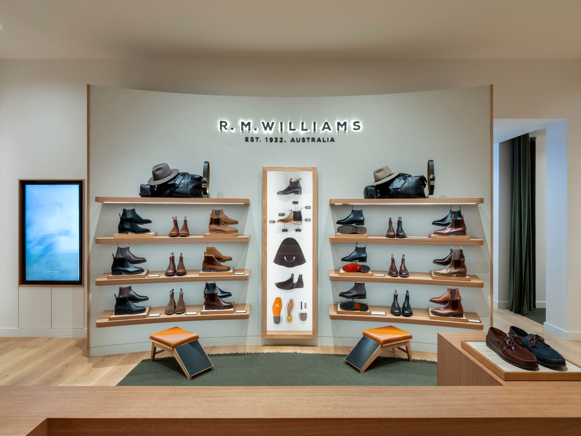R.M.Williams store display with boots, hats, and leather goods on shelves against a beige wall. A digital screen and wooden stools are visible.