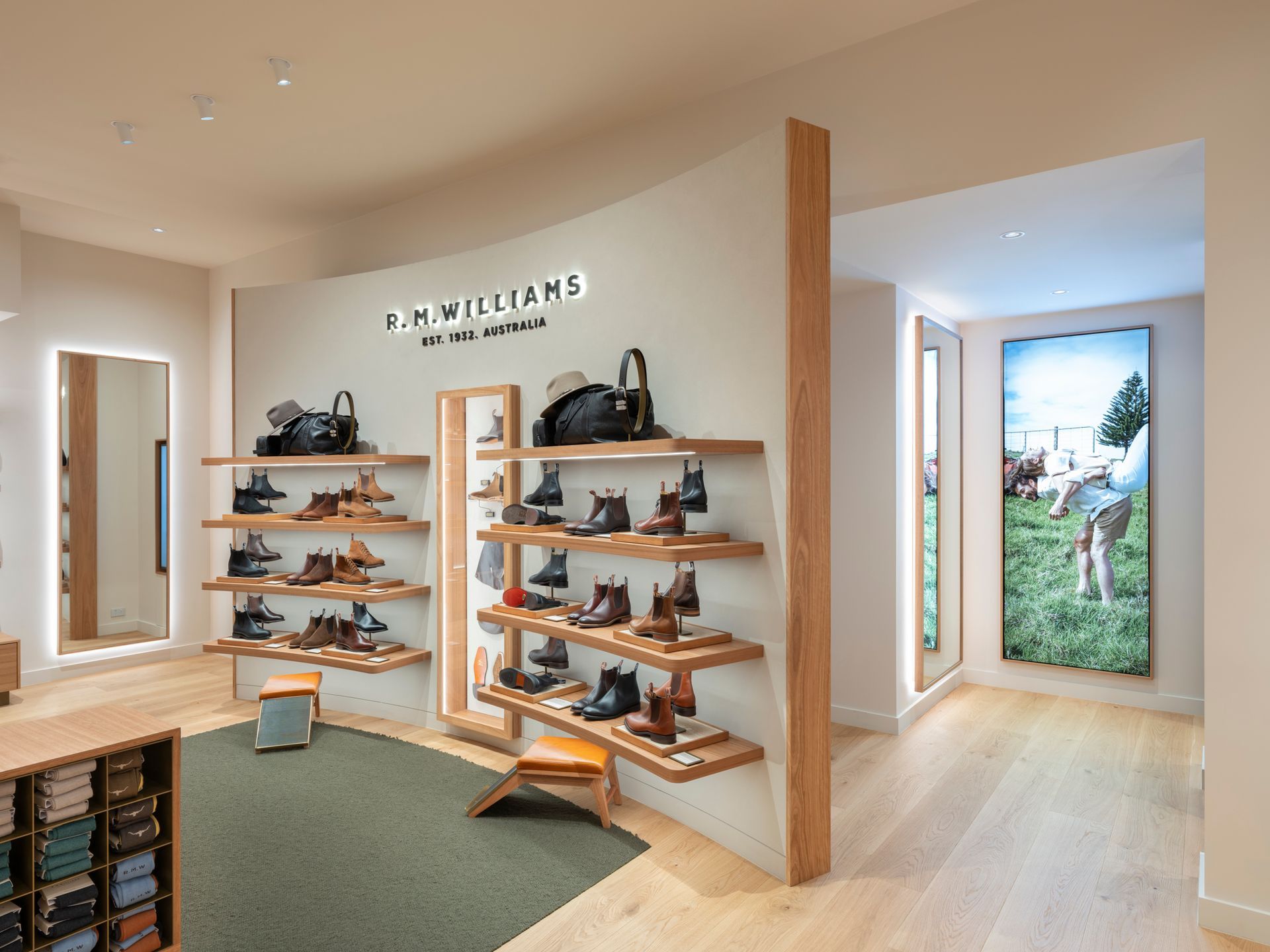 Interior of a R.M. Williams store displaying boots on wooden shelves. Features neutral color palette, hardwood floors, and a curved wall.