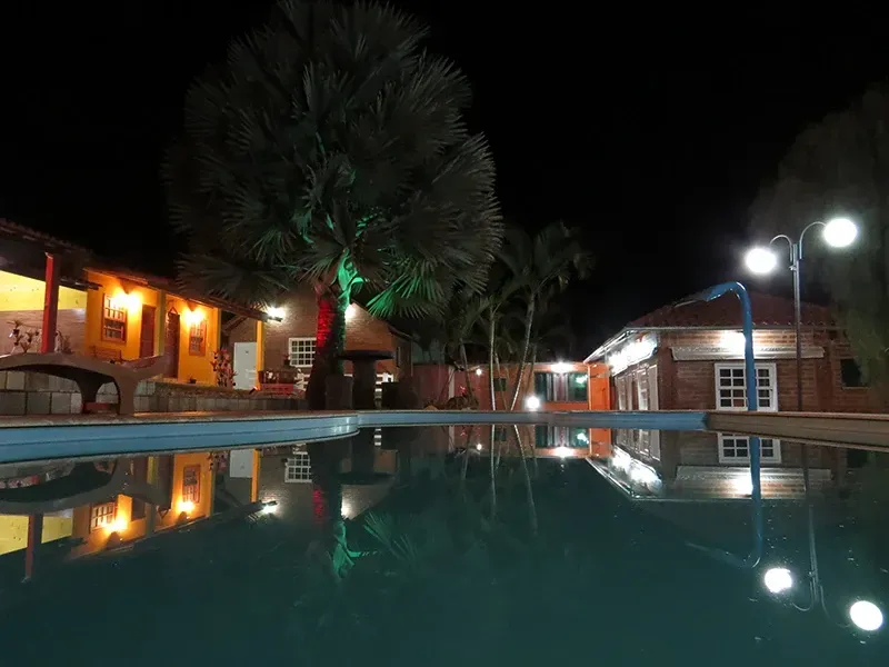 Vista noturna de edifícios e palmeiras refletidos em um lago; luzes alaranjadas iluminam a cena.