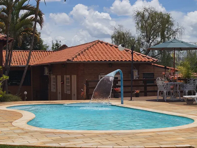Piscina com fonte em frente a um prédio de tijolos com telhado laranja. Dia ensolarado.