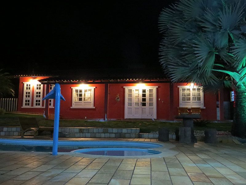 Prédio vermelho com detalhes brancos e janelas iluminadas à noite, próximo a uma piscina e uma palmeira.