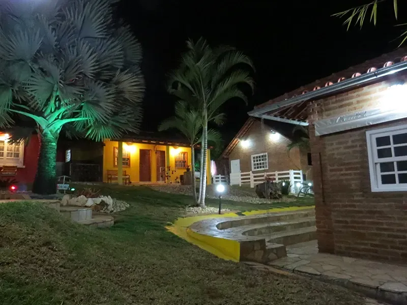 Vista noturna de edifícios iluminados e palmeiras em uma colina gramada; um caminho e degraus levam às estruturas.
