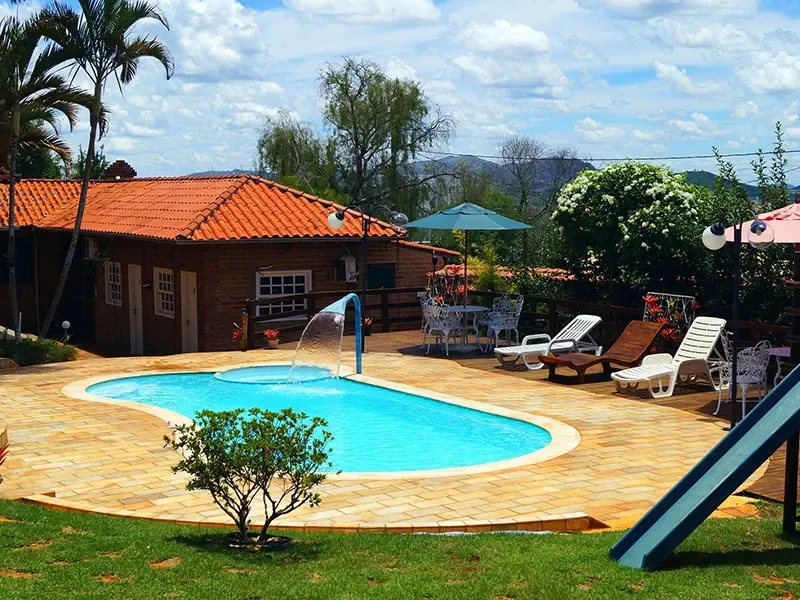 Área da piscina com uma pequena construção, espreguiçadeiras e um escorregador; ambiente externo ensolarado.
