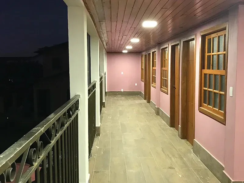 Corredor externo com paredes rosa, portas e janelas de madeira e um corrimão escuro.