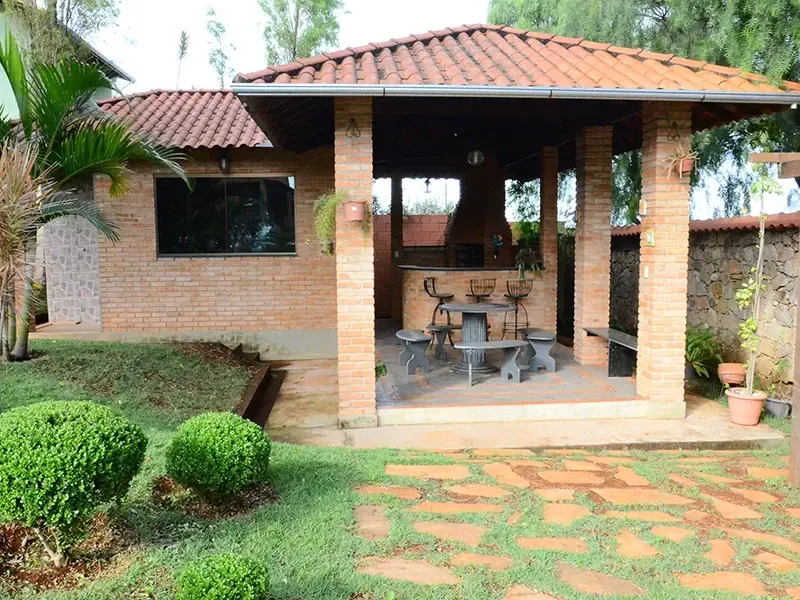 Pátio de tijolos com telhado de telhas, aberto para o quintal, com mesa e churrasqueira.