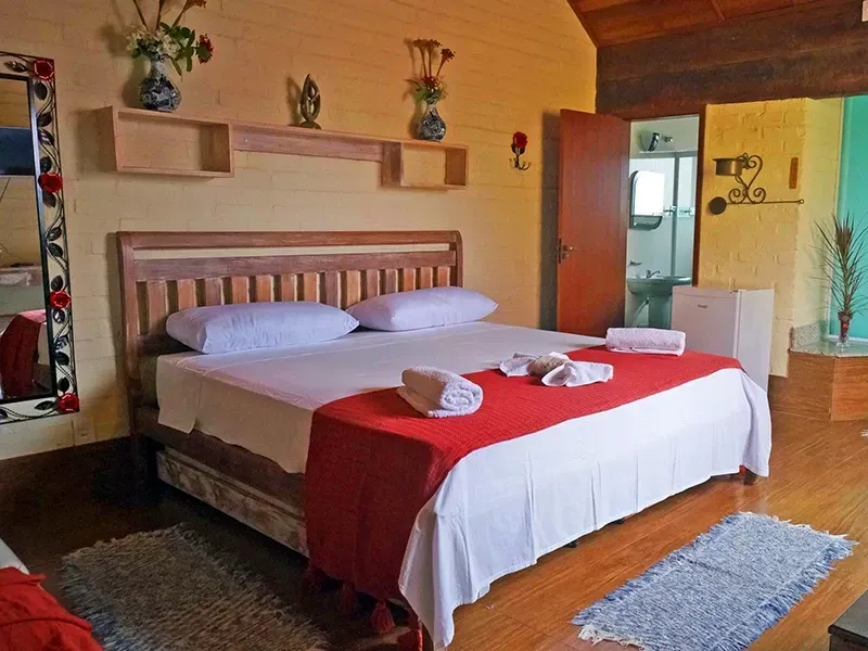 Quarto com cama, cobertor vermelho, móveis de madeira, vasos e porta aberta para o banheiro.