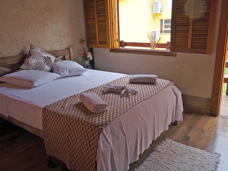 Quarto com cama de madeira, roupa de cama branca e peseira decorativa. Uma janela com persianas está aberta.