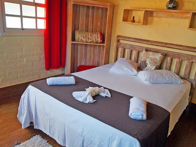 Quarto com cama, lençóis brancos, passadeira marrom, cortinas vermelhas e móveis de madeira.