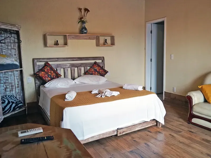 Quarto com cama de casal, sofá, móveis de madeira e porta. Interior ensolarado.