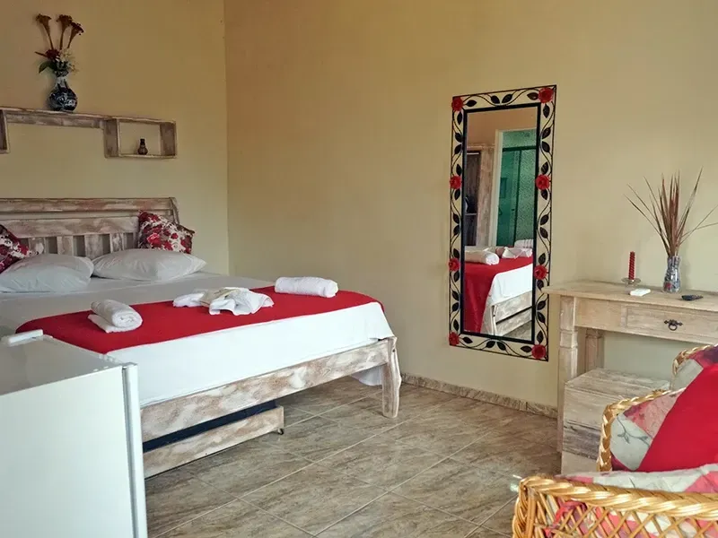 Quarto com cama branca, detalhes em vermelho, um grande espelho e uma pequena mesa de madeira.