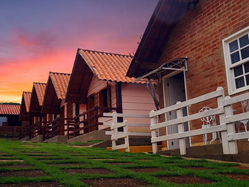 Fileira de pequenas cabanas de tijolo e madeira com cercas brancas, tendo como pano de fundo o céu ao pôr do sol.