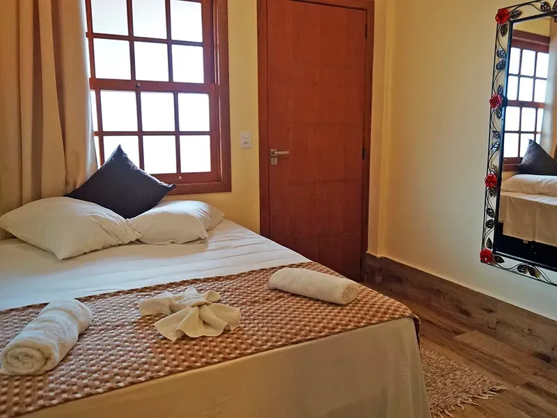 Quarto com cama, toalhas, janela e espelho. Tons de bege e marrom.