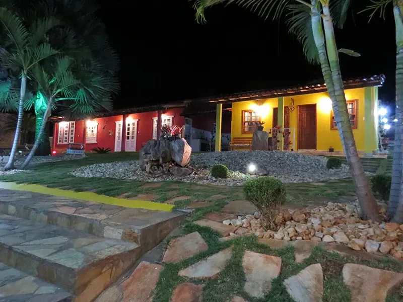 Edifícios coloridos com paredes vermelhas e amarelas, à noite, rodeados por jardins e palmeiras iluminadas.