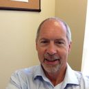 A middle-aged man with graying hair and a goatee smiles, wearing a light blue shirt, in an office setting.