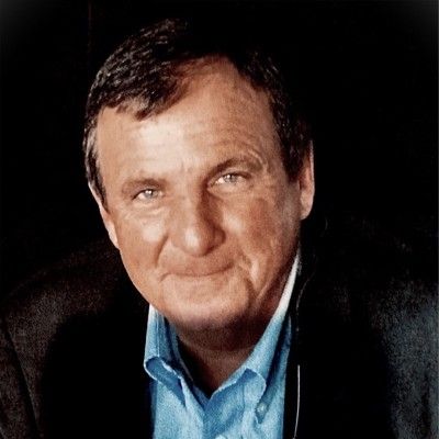 Smiling older man with fair skin, blue eyes, and dark hair wearing a blue shirt and black jacket.