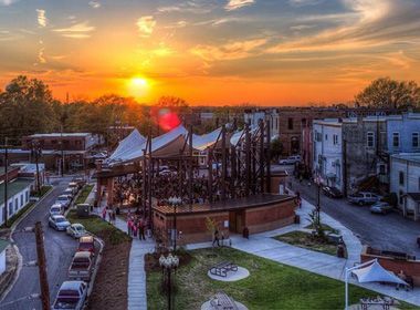 Sunset over a town square with people, buildings, and an art installation. Cars line the streets.