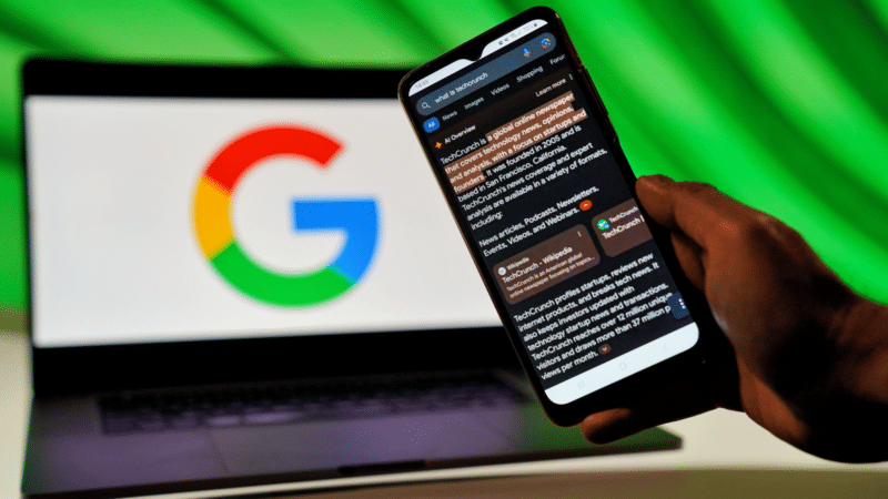 Person holding a smartphone with a Twitter feed, Google logo in the background on a laptop. Green backdrop.