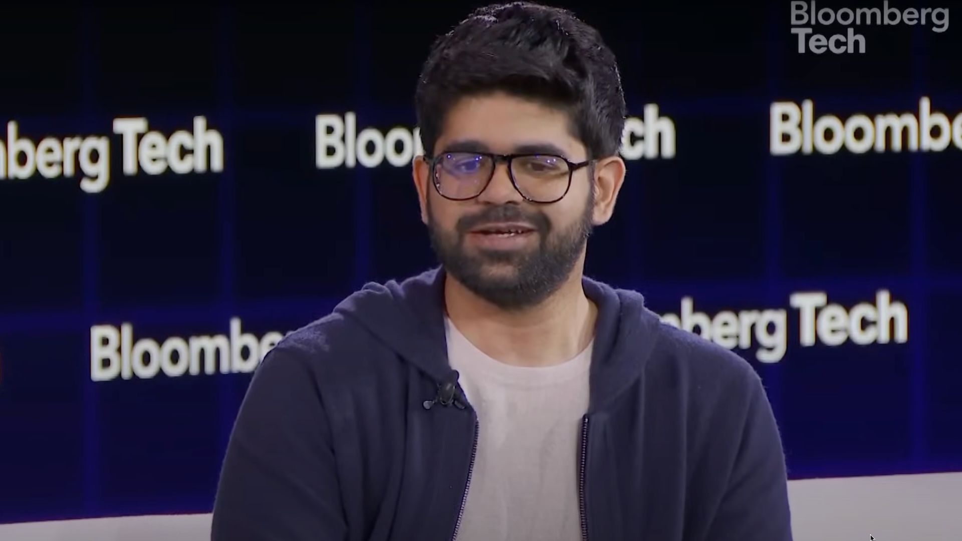 Man with glasses in a blue hoodie talking at a Bloomberg Tech event.