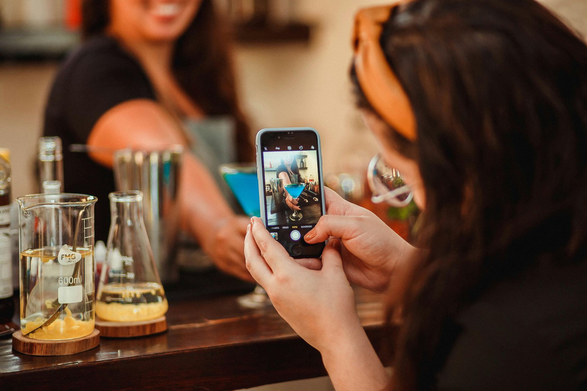 Woman taking photo with phone of another woman behind a bar mixing drinks.