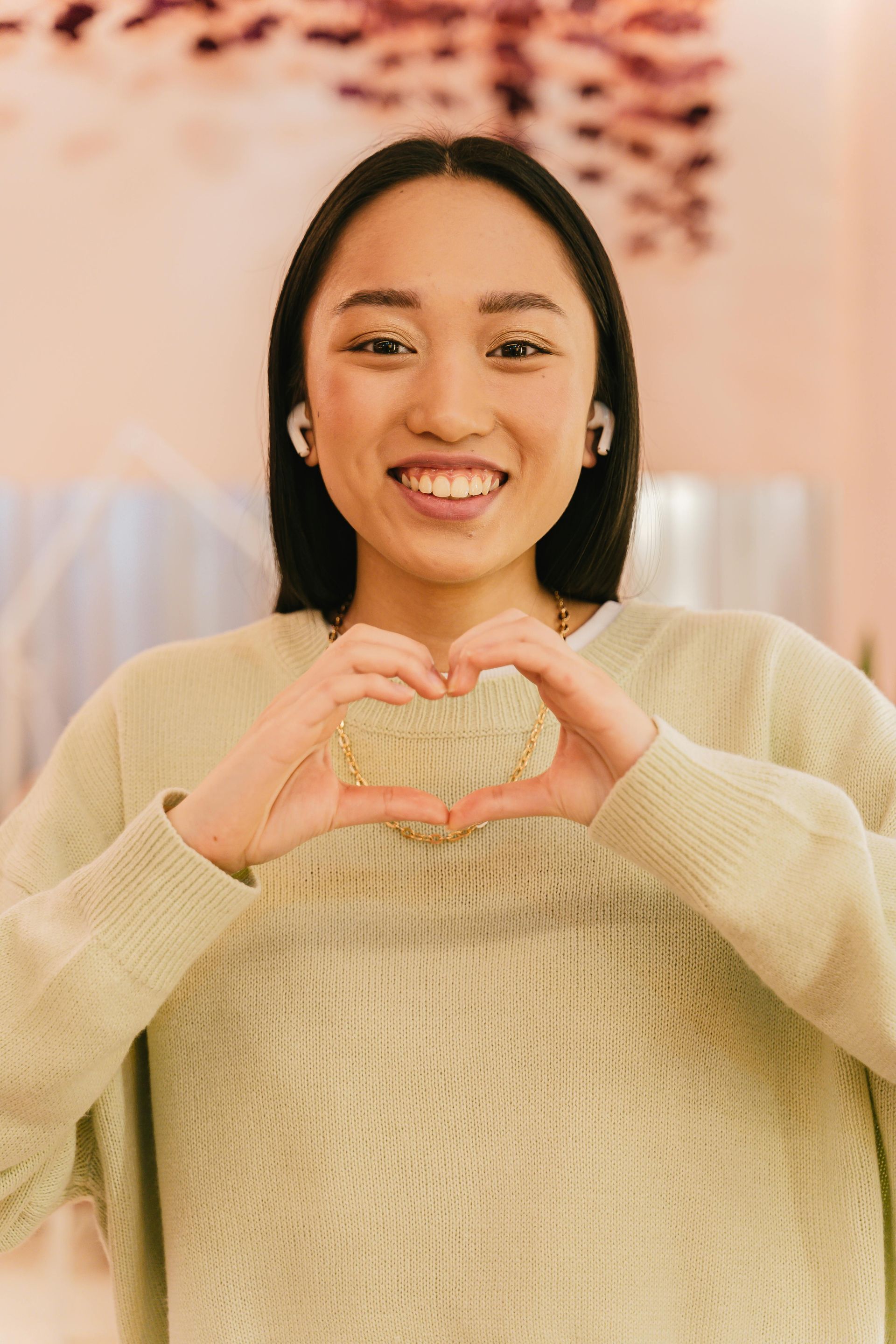 A young Asian woman with long black hair makes a heart shape with her hands. She smiles widely while wearing a light green sweater.