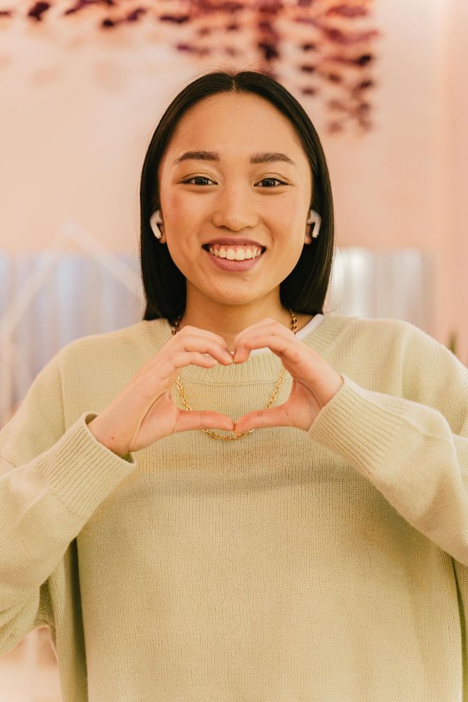 A young Asian woman with long black hair makes a heart shape with her hands. She smiles widely while wearing a light green sweater.