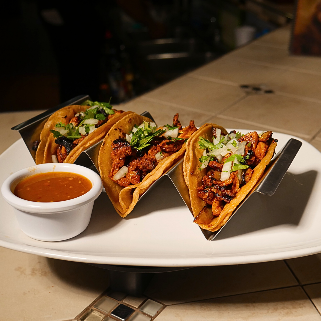 Three tacos with meat and cilantro on a plate, with a bowl of sauce next to them.