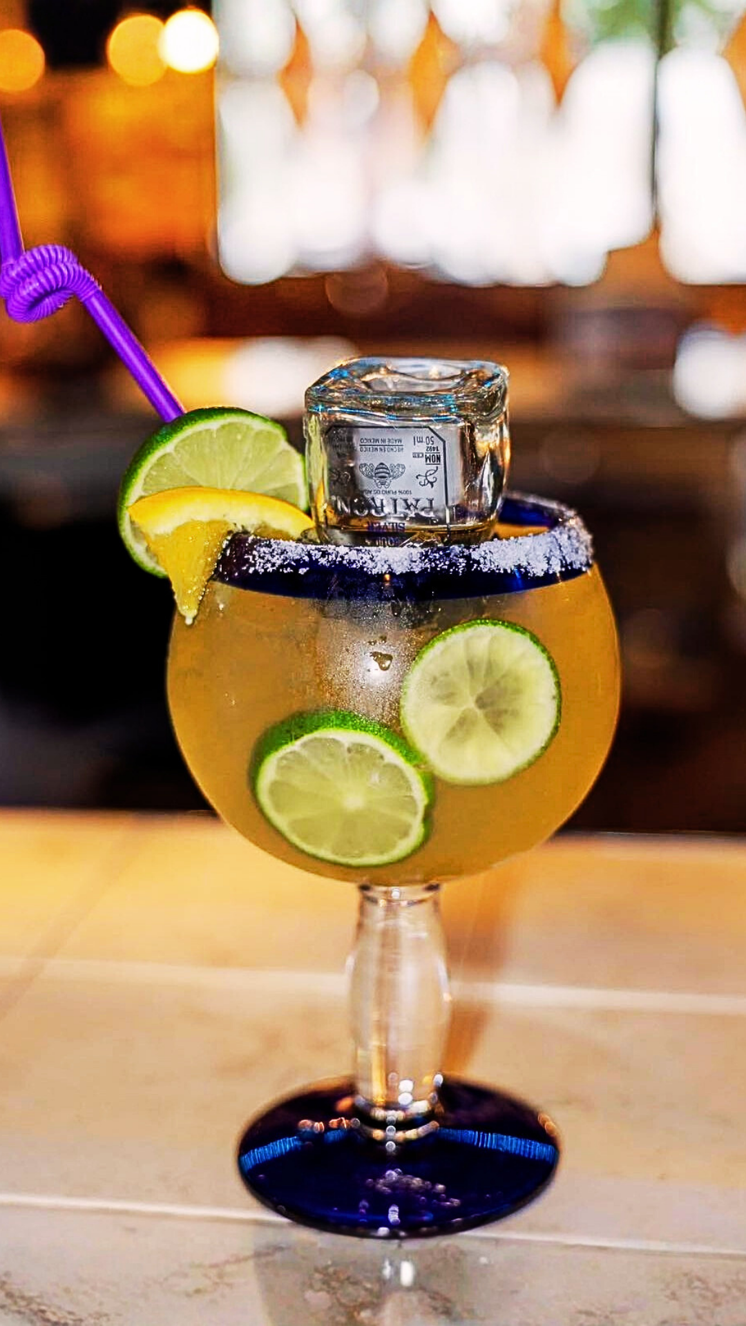 Large margarita glass with lime garnish, salt rim, shot of tequila, purple straw.