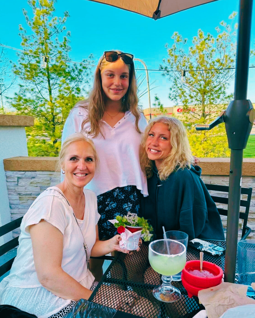 Three people at outdoor table; woman holding gift, two younger people smile. Drinks, umbrella, and greenery in background.