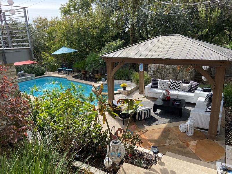 Backyard with pool, gazebo with white seating, blue umbrella, and lush greenery.