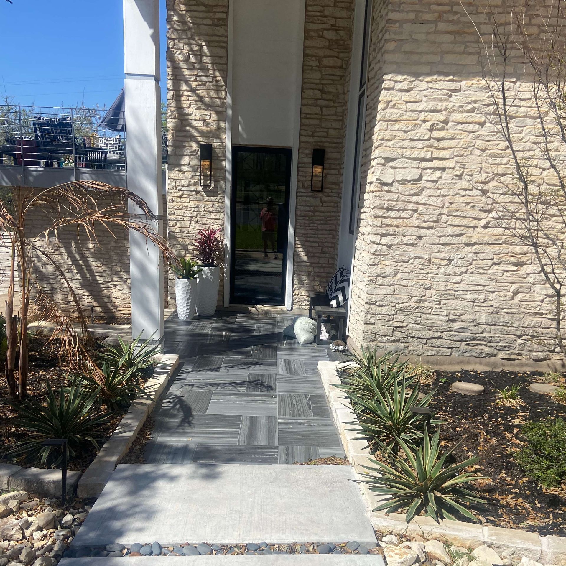 Entrance of a modern home with stone facade, patterned walkway, plants, and black front door.