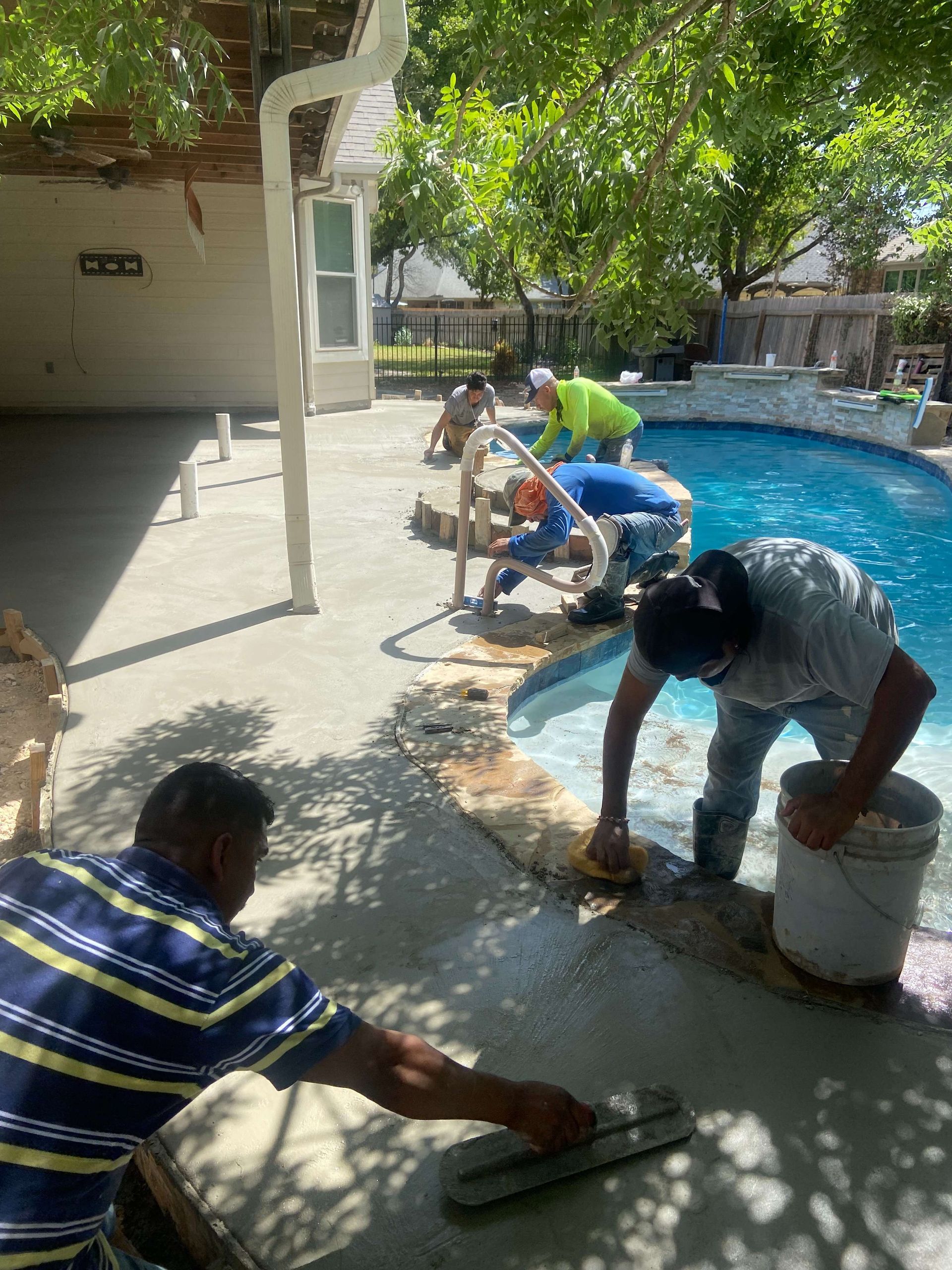 Men resurfacing a pool deck with concrete. Blue pool water, green trees, sunny day.