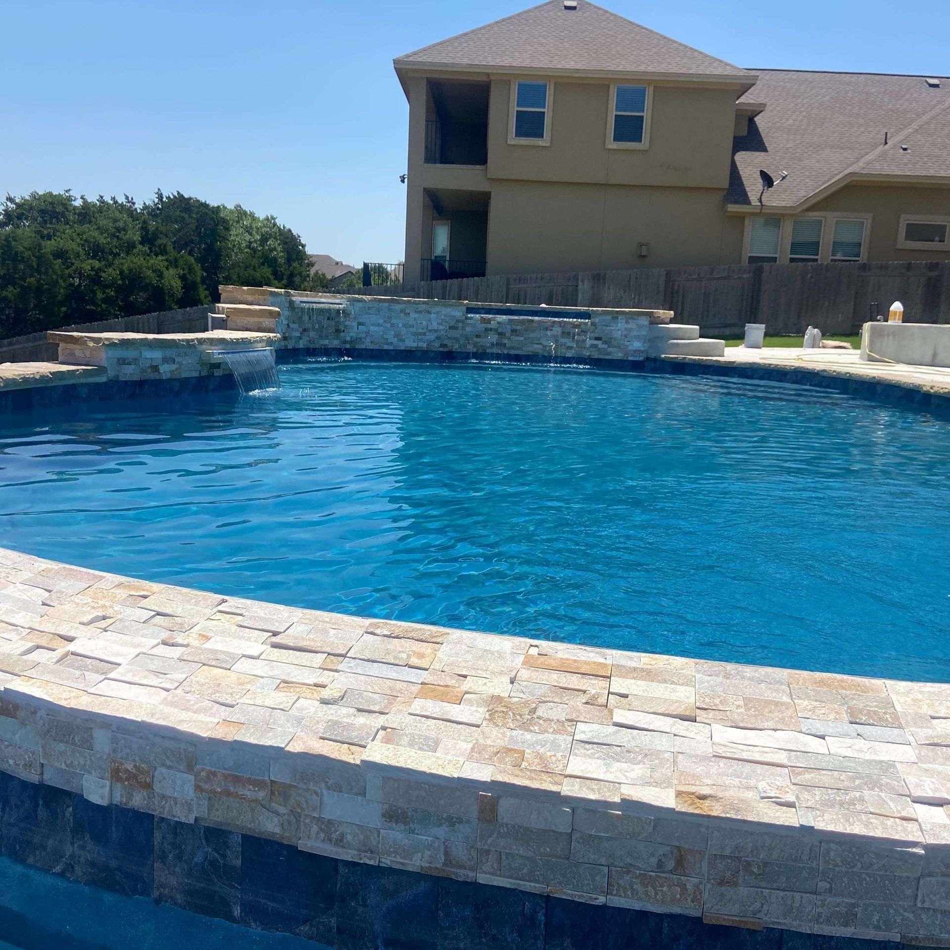 Swimming pool with stone edging, blue water, and a beige house in the background on a sunny day.