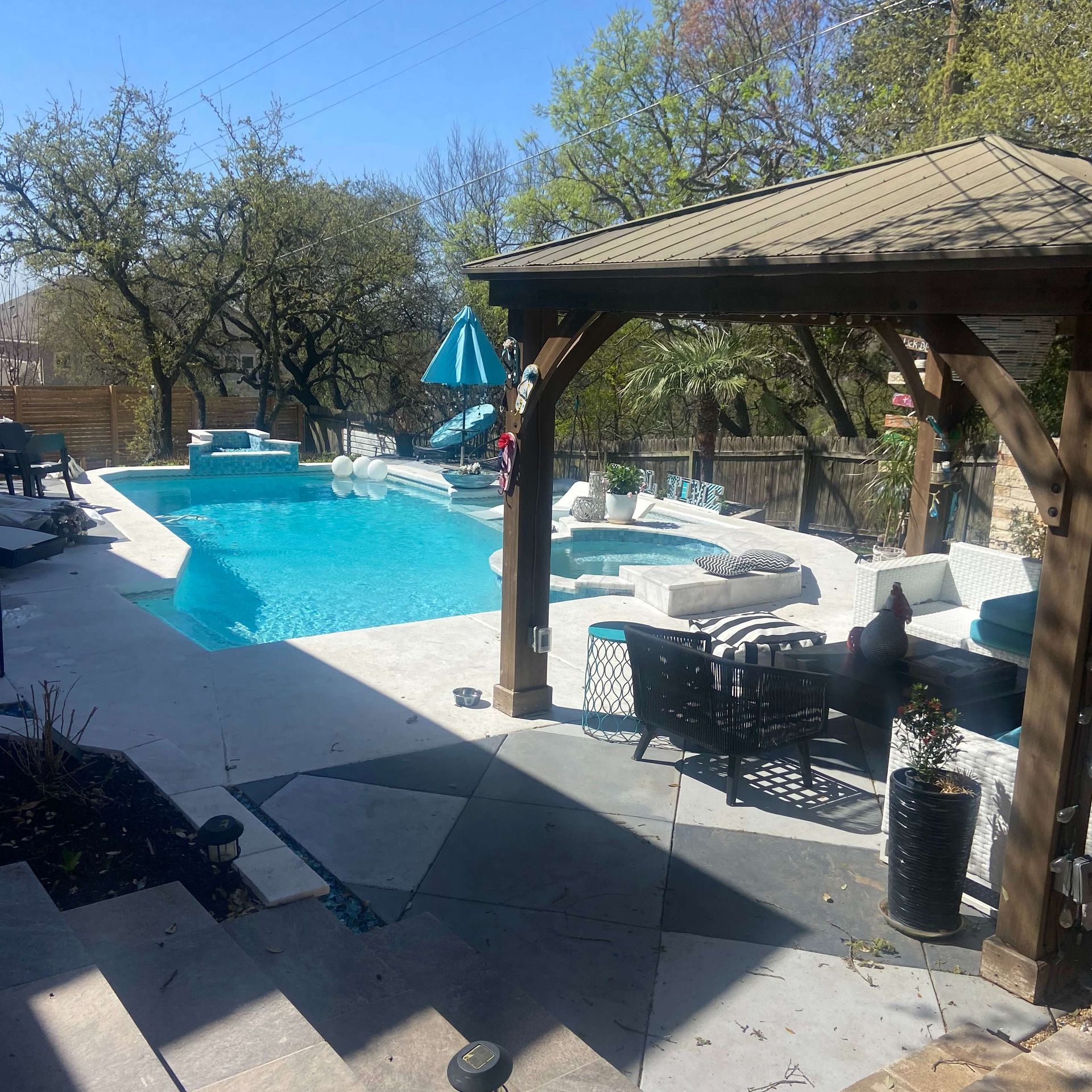 Backyard pool with gazebo, seating, and a blue umbrella. Bright, sunny day.