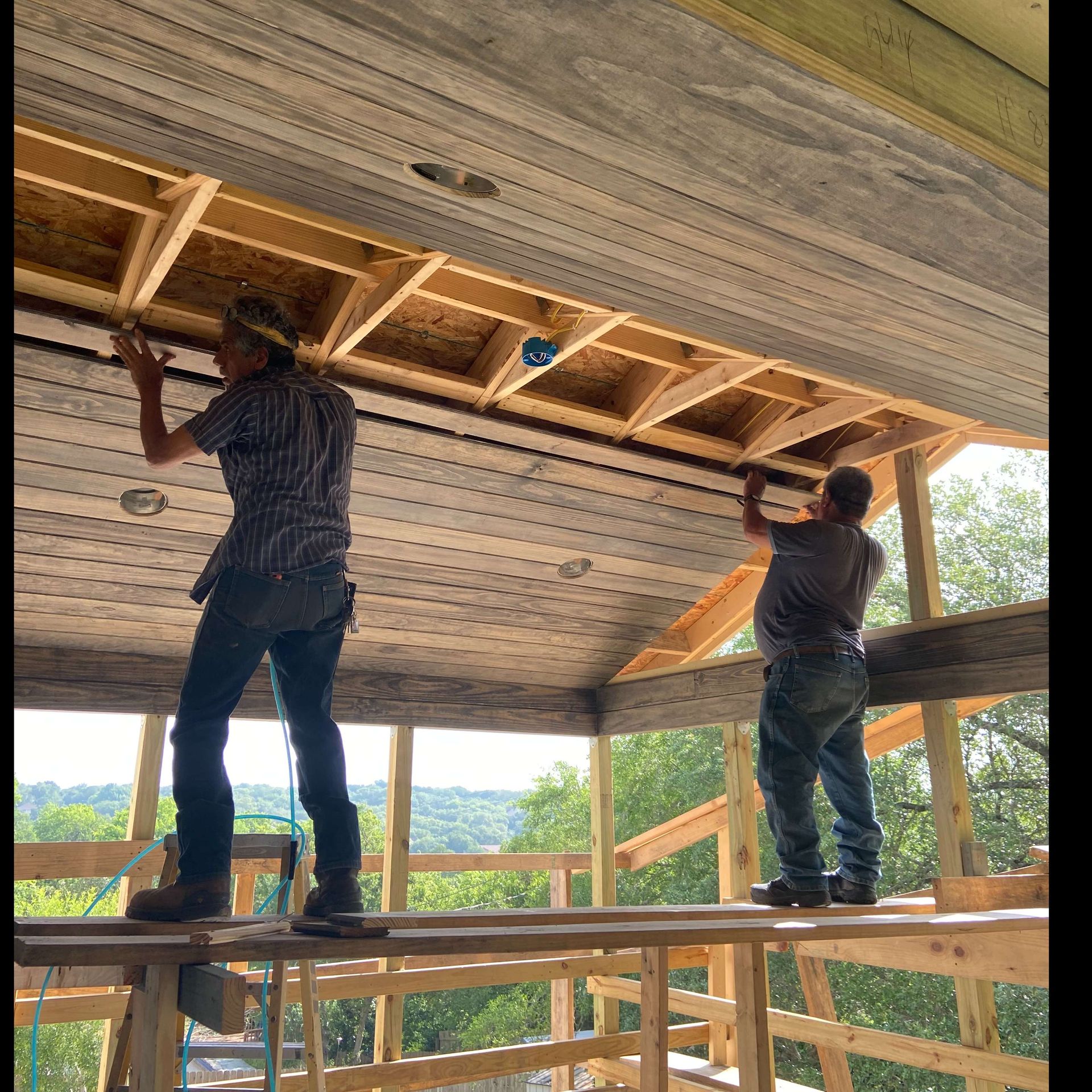 Two construction workers install a ceiling on a wood-framed structure outdoors.
