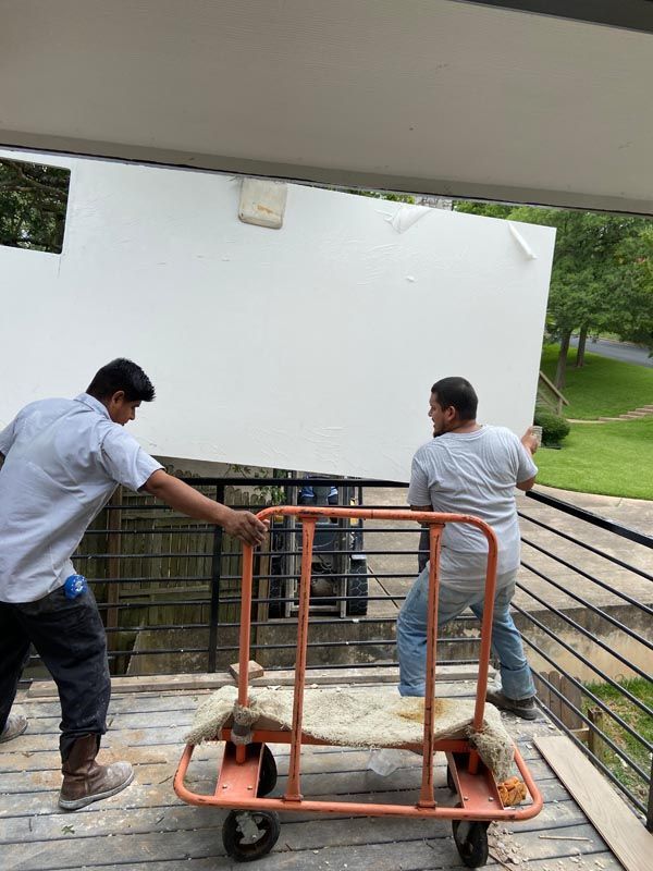 Two men lift a large white panel onto a hand truck on a balcony.