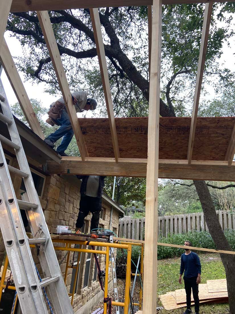 Construction workers building a roof extension; one on the roof, one on scaffolding, and another standing below.