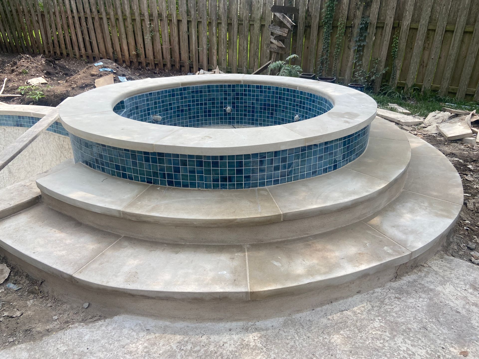 Circular hot tub with blue tile, two stone steps, and a concrete patio in a yard.