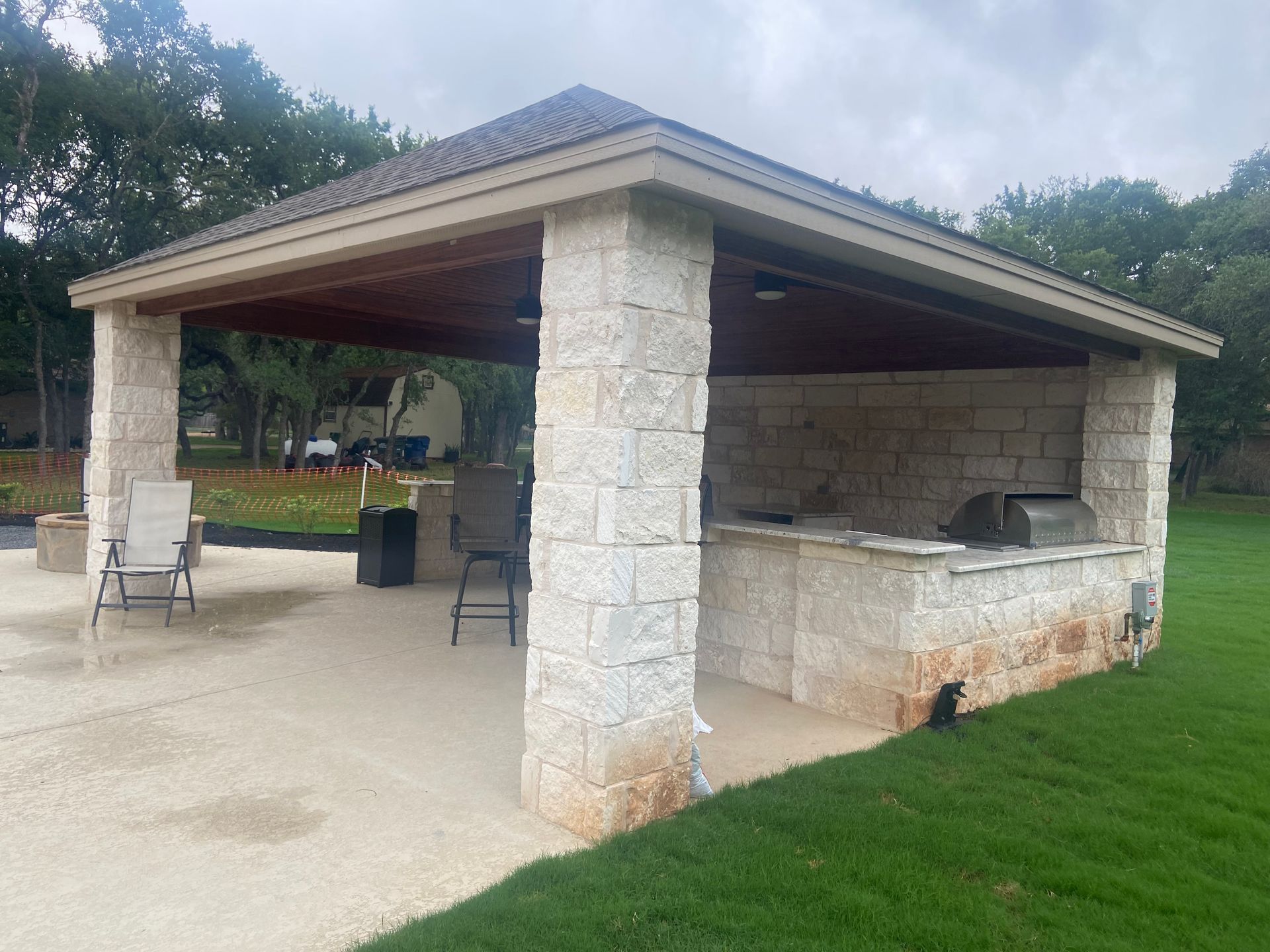 Stone-columned outdoor kitchen under a brown roof on a concrete patio, with chairs and a grill.