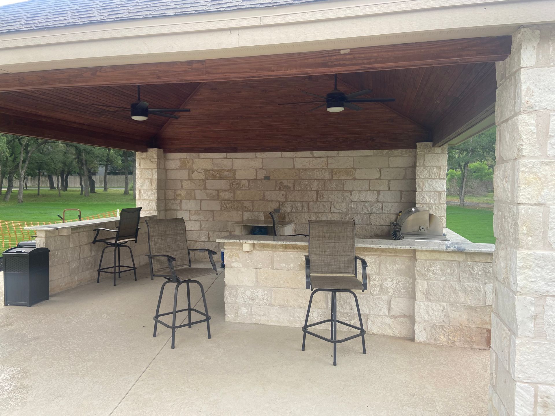 Outdoor stone bar with stools under a covered patio, two ceiling fans, grassy background.