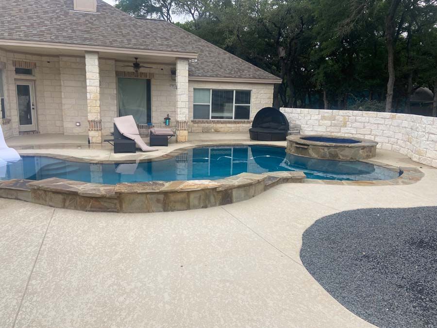 Pool and hot tub in a backyard with light-colored house and stone wall.