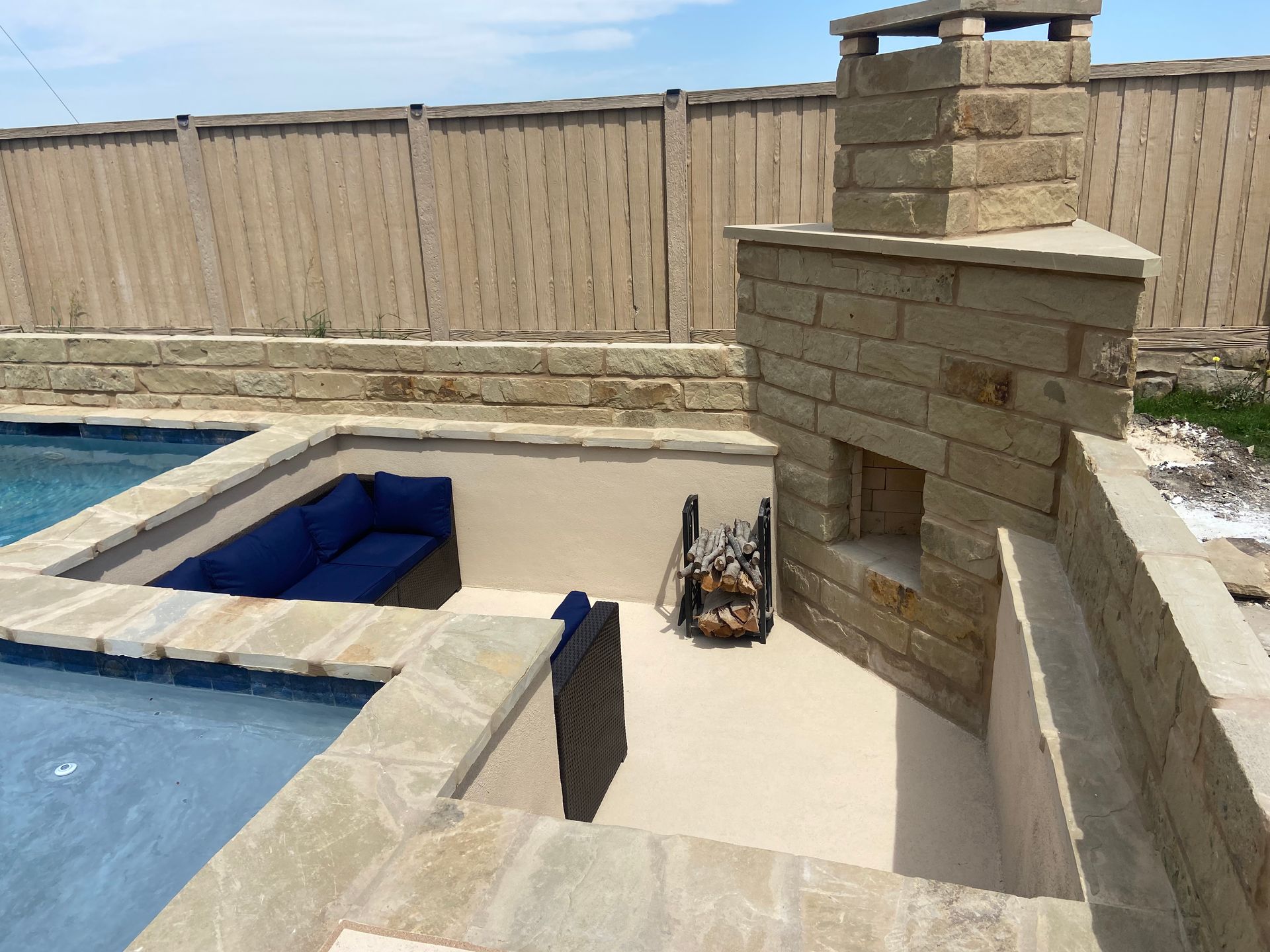 Backyard pool with a stone fireplace and seating area. Blue pool water, beige stone, blue couch.