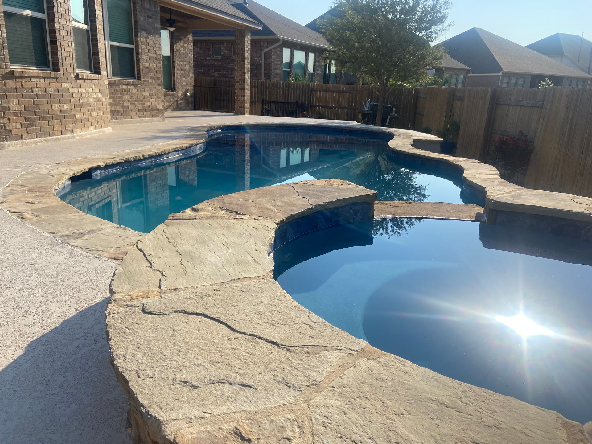 Pool with stone coping, blue water, surrounded by patio, house in background.