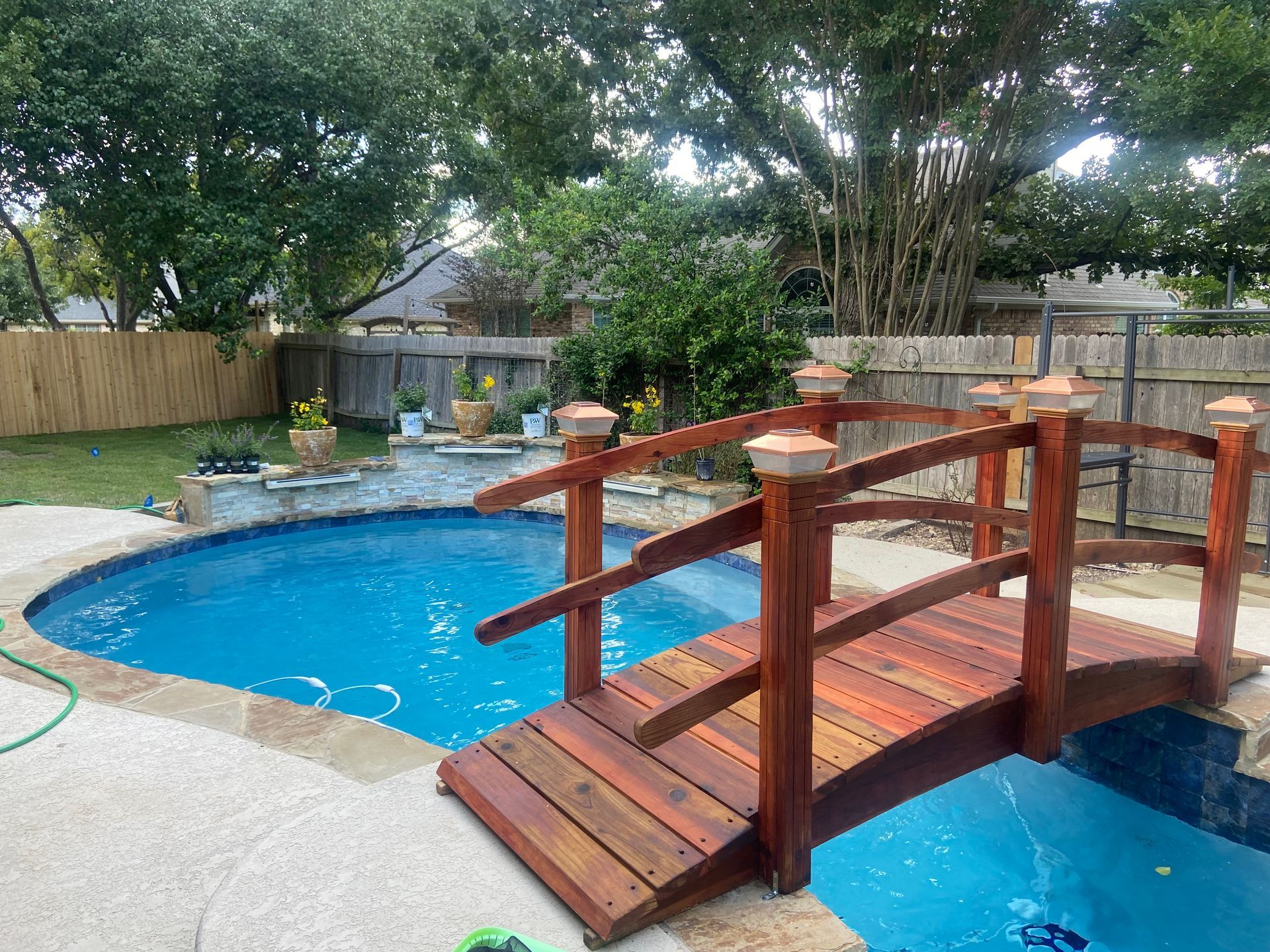 Pool with a wooden bridge, surrounded by trees and a stone patio.