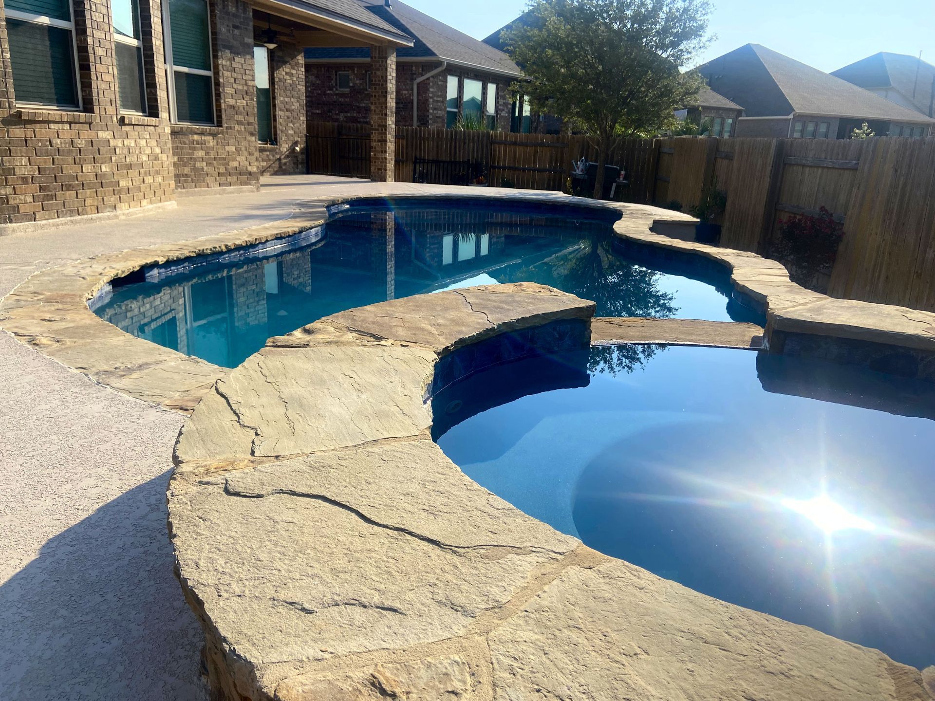 Pool with stone coping and clear blue water, surrounded by a patio and fencing, on a sunny day.