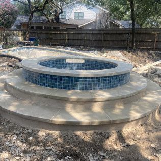 Circular in-ground spa with blue tile and tan steps, under construction in a backyard.