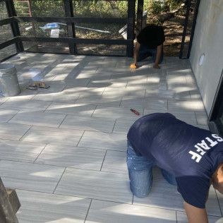 Two workers tiling a gray-tiled patio. One spreads grout, the other kneels. Sunny outdoor setting.