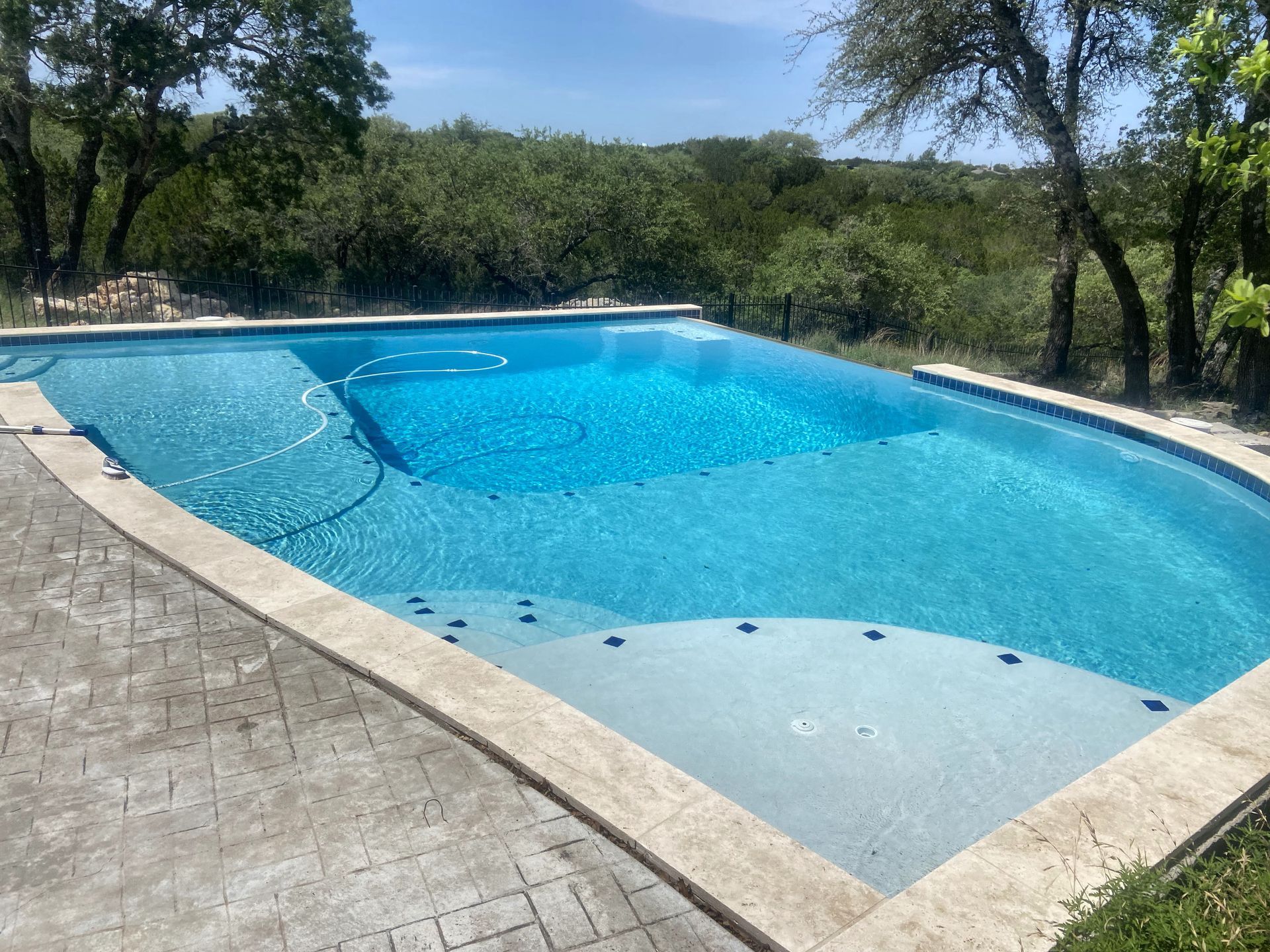 A rectangular blue pool with a step-in area, surrounded by a stone patio, set against a green landscape.