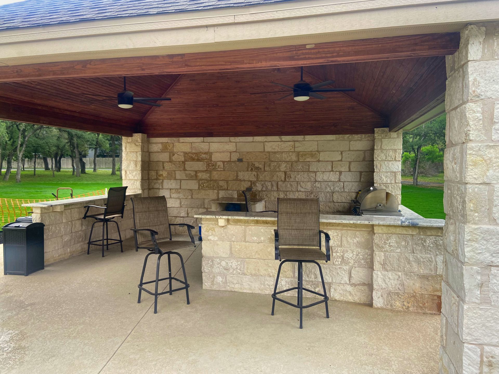 Outdoor kitchen with stone walls, bar seating, and ceiling fans.