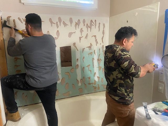 Two men removing wall tiles from a bathtub surround with a hammer and a power tool.