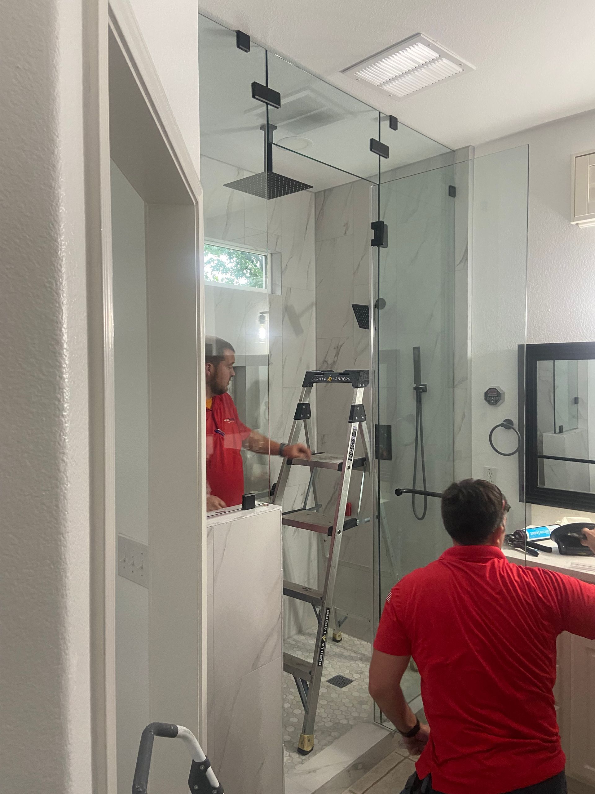 Two men in red shirts installing a frameless glass shower in a bathroom. One on a ladder.